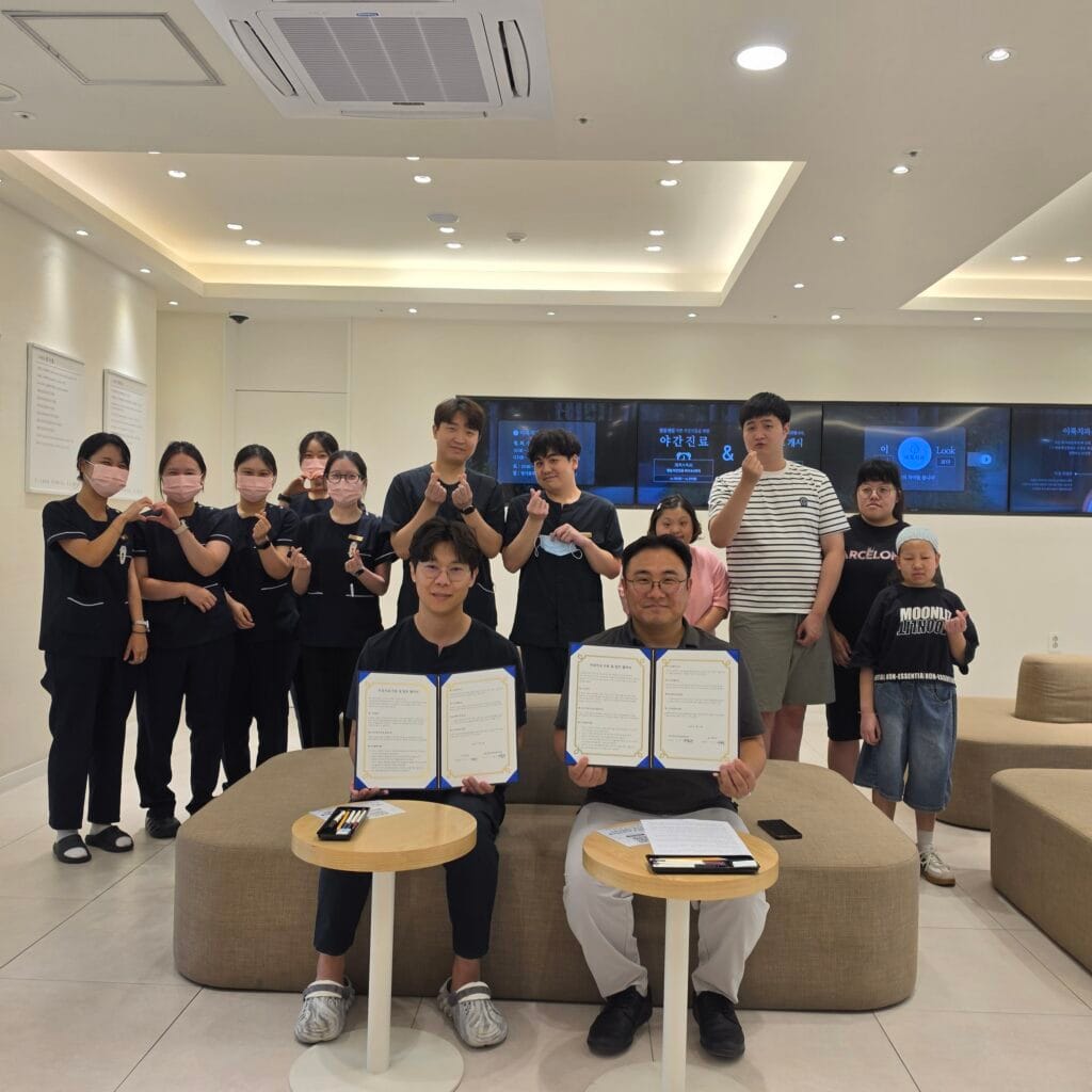 MOU signing ceremony at 365 E-LOOK Dental's lobby. Dr. Junsang Lee and Eumteo center director Jongchan Lee hold the signed agreement, surrounded by clinic staff and Eumteo trainees making finger hearts for a group photo.